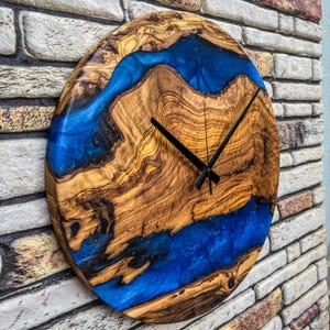 Puede incluir: Un reloj de pared redondo de madera con un dise&ntilde;o &uacute;nico. La esfera del reloj presenta un patr&oacute;n de vetas de madera natural con un r&iacute;o de resina azul vibrante. Las manecillas negras indican la hora. El reloj est&aacute; montado en una pared de ladrillo.