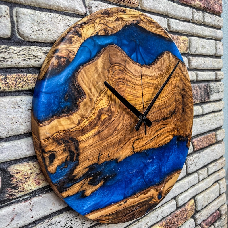 Puede incluir: Un reloj de pared redondo de madera con un dise&ntilde;o &uacute;nico. La esfera del reloj presenta un patr&oacute;n de vetas de madera natural con un r&iacute;o de resina azul vibrante. Las manecillas negras indican la hora. El reloj est&aacute; montado en una pared de ladrillo.