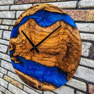 Puede incluir: Un reloj de pared redondo de madera con un dise&ntilde;o llamativo. La esfera presenta un patr&oacute;n de vetas de madera natural con un r&iacute;o de resina azul vibrante. Las manecillas negras indican la hora. Di&aacute;metro aproximado de 30 cm.