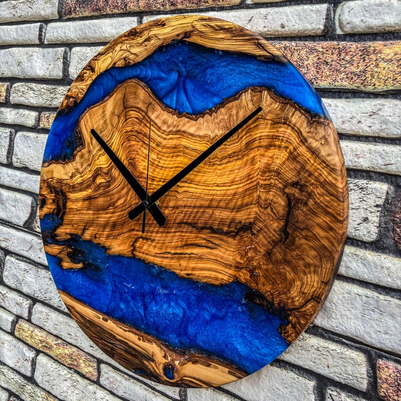 Puede incluir: Un reloj de pared redondo de madera con un dise&ntilde;o llamativo. La esfera presenta un patr&oacute;n de vetas de madera natural con un r&iacute;o de resina azul vibrante. Las manecillas negras indican la hora. Di&aacute;metro aproximado de 30 cm.