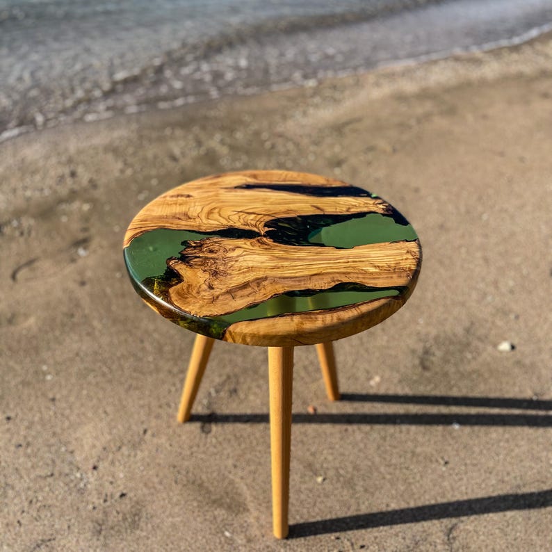 May include: A round wooden table with a green and black epoxy resin top. The table has three wooden legs and is sitting on a sandy beach.