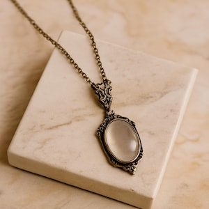 May include: An antique-style necklace with an oval pendant. The pendant has a light-colored, oval-shaped stone set in an ornate, bronze-colored frame. The chain is also bronze-colored and hangs from the pendant.