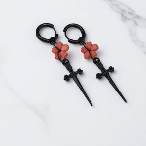 Puede incluir: Un par de pendientes de aro negros con un diseño colgante. Cada pendiente presenta una pequeña flor de color coral y un colgante de daga negra. Los pendientes se muestran sobre una superficie de mármol blanco.