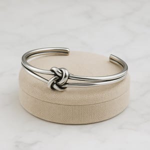 May include: A silver-coloured cuff bracelet with a knot design. The bracelet has a double-band design that wraps around the wrist, with a knot detail at the centre. The bracelet is displayed on a beige, circular surface.