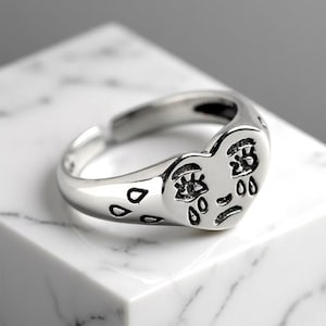 Puede incluir: Un anillo plateado con un diseño de cara en forma de corazón. La cara tiene ojos, boca y lágrimas. El anillo está sobre una superficie de mármol blanco.