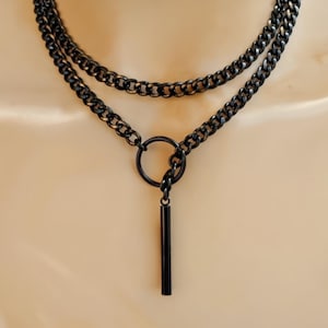 Könnte beinhalten: Eine schwarze Halskette mit einem mehrschichtigen Kettendesign. Die Halskette hat einen kreisförmigen Anhänger und einen vertikalen Stangenanhänger. Die Kette ist aus Metall und hat ein glänzendes Finish. Die Halskette wird vor einem neutralen Hintergrund präsentiert.