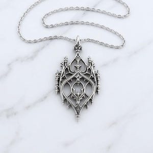 Pode incluir: Um colar com pingente de metal prateado apresentando um design vazado intrincado e simétrico com elementos pontiagudos em forma de agulha e linhas curvas. O pingente está suspenso por uma corrente a condizer com elos ovais.