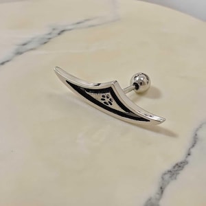 May include: A silver-colored ear piercing with a curved, blade-like design. The piercing features a small, decorative black pattern and a ball-shaped backing. The jewelry is designed to be worn in the ear.