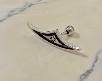 May include: A silver ear piercing with a curved, blade-like design. The piercing has a small, decorative black pattern and a ball-shaped backing. The jewellery is designed to be worn in the ear.