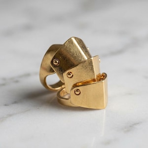 May include: A gold-toned articulated ring featuring three overlapping shield-like plates connected by small rivets. The metal surface has a brushed finish, and the design consists of a main band with two additional hinged segments that extend outward.