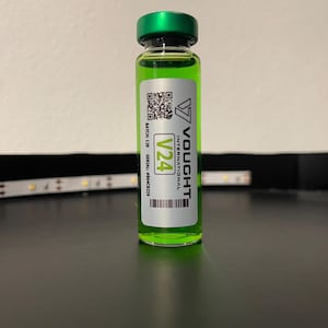 Può includere: Una piccola fiala di vetro riempita con un liquido verde brillante, sigillata con un tappo verde. La fiala ha un'etichetta bianca con il testo "Vought International" e "V24". L'etichetta include anche un codice QR e un codice a barre.