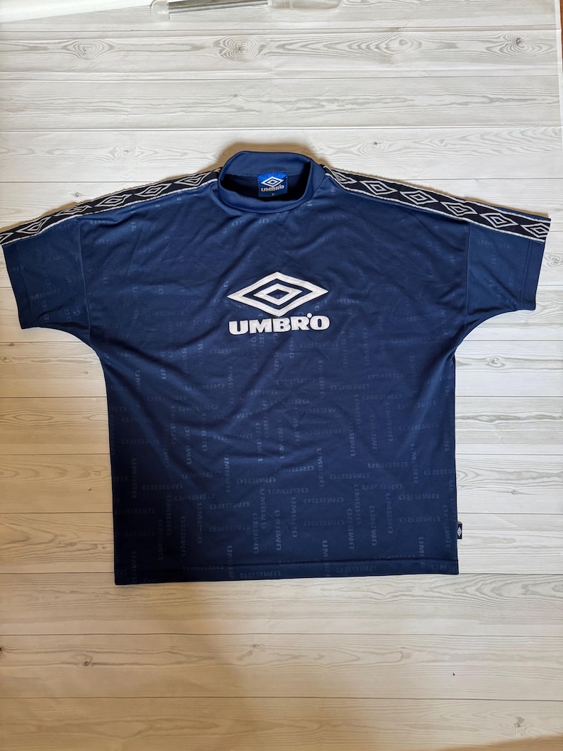 May include: Navy blue Umbro soccer jersey with a mock neck collar. The jersey features the Umbro logo in white on the chest and repeating text. White and black geometric pattern on the shoulders.