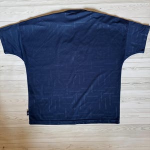 May include: Navy blue short-sleeve shirt with a subtle all-over pattern. The shirt has a crew neck and a relaxed fit. The fabric appears to be a lightweight material, suitable for casual wear. The brand name is repeated throughout the pattern.