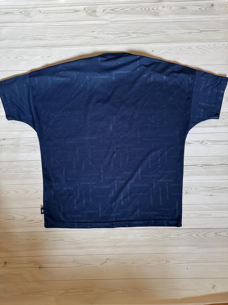 May include: Navy blue short-sleeve shirt with a subtle all-over pattern. The shirt has a crew neck and a relaxed fit. The fabric appears to be a lightweight material, suitable for casual wear. The brand name is repeated throughout the pattern.