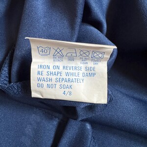 May include: A close-up of a navy blue fabric with a white care label attached. The label displays washing instructions, including a 40&deg; wash, do not bleach, cool iron, do not dry clean, and do not tumble dry. The label also says to iron on the reverse side, reshape while damp, wash separately, and do not soak.