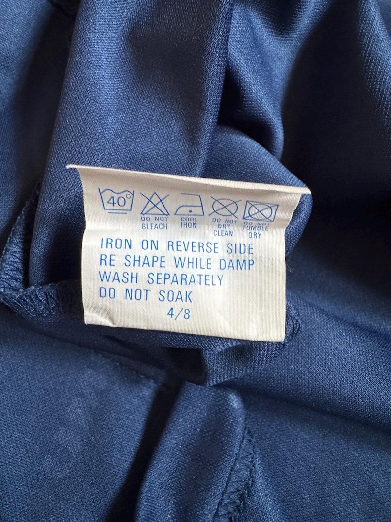 May include: A close-up of a navy blue fabric with a white care label attached. The label displays washing instructions, including a 40&deg; wash, do not bleach, cool iron, do not dry clean, and do not tumble dry. The label also says to iron on the reverse side, reshape while damp, wash separately, and do not soak.