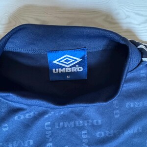 May include: A navy blue athletic shirt with the Umbro logo on the tag. The shirt has a repeating Umbro pattern across the fabric. The tag also has the size "M" printed on it.