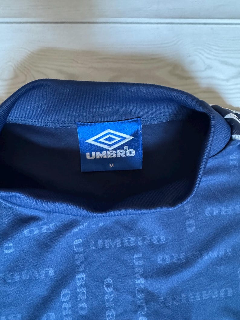 May include: A navy blue athletic shirt with the Umbro logo on the tag. The shirt has a repeating Umbro pattern across the fabric. The tag also has the size "M" printed on it.