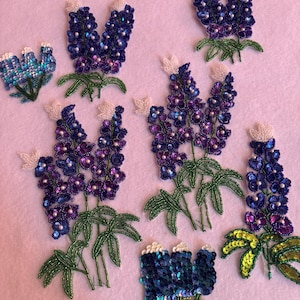 May include: A collection of seven bluebonnet flower appliques, each made with sequins and beads. The appliques are in various sizes and feature different shades of blue and purple.