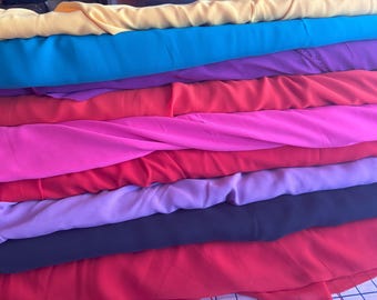 May include: A stack of colorful fabric rolls in shades of yellow, teal, purple, red, pink, and black. The fabric appears to be a lightweight material, possibly chiffon or silk, with a slight sheen. The rolls are neatly stacked, showcasing the variety of colors.