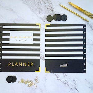May include: A black and white striped planner with gold accents, open to display the interior. The planner includes the text "PLANS TO SUCCEED" and "PLANNER". Gold scissors, a pen, paper clips, and black discs are also visible.