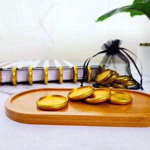 May include: A wooden tray holds several gold-colored round objects. A notebook with gold rings and a black and white striped cover is in the background. A small black bag and a potted plant are also visible.