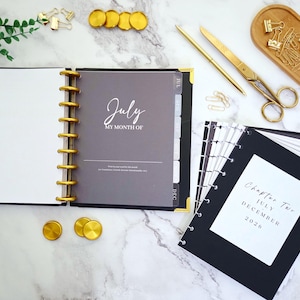 May include: An open black and gold planner with the text "July My Month Of" on a gray page. Gold accessories include a pen, scissors, paper clips, and decorative coins. The planner is on a marble surface.