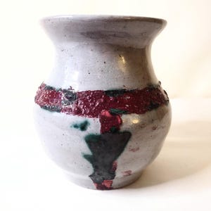 May include: A handcrafted ceramic vase with a light gray body and a textured, dark red band around the middle. The vase features a flared rim and a decorative black and green design. This unique vase is perfect for displaying flowers or as a decorative accent.