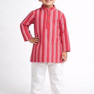 Puede incluir: Un conjunto infantil que incluye una camisa kurta a rayas rojas y blancas con cuello mandarín y botones de madera, combinada con pantalones blancos. La camisa kurta tiene mangas largas. El atuendo es adecuado para una ocasión festiva.