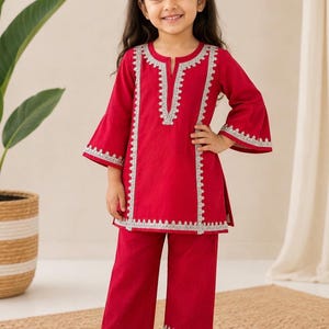 May include: A vibrant red two-piece outfit consisting of a long-sleeved tunic with a notched neckline and wide-leg trousers. The tunic features intricate silver embroidery along the neckline, front panels, and sleeve cuffs, with matching silver trim on the hem of the trousers.