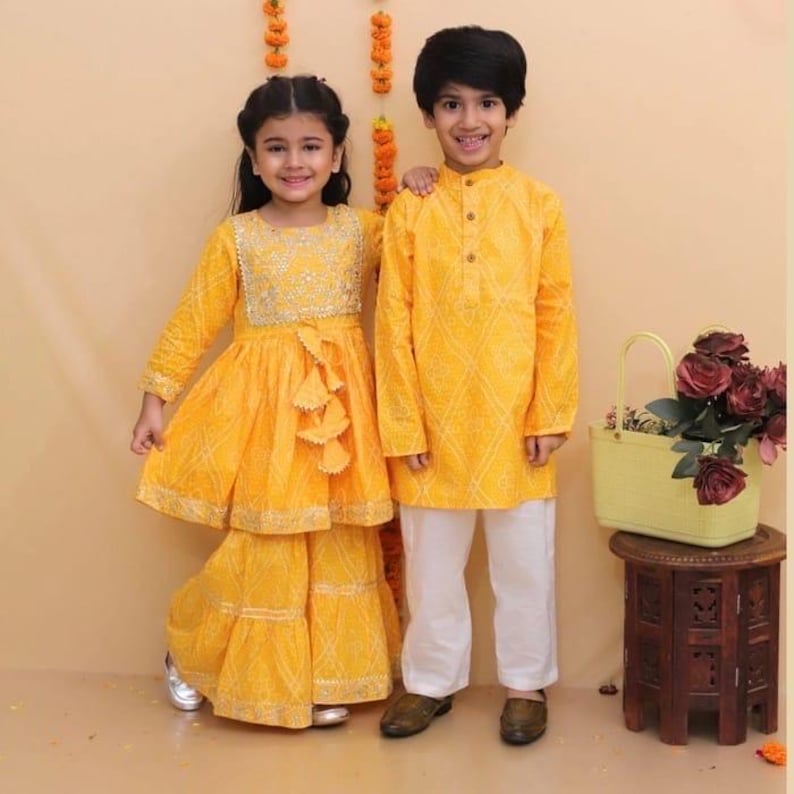 May include: A yellow patterned peplum top with a matching tiered skirt featuring silver embroidery and tassels, and a yellow long-sleeved tunic paired with plain white trousers. A light yellow basket containing dark red roses sits nearby.