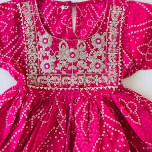 May include: A bright pink dress featuring a white tie-dye pattern and a square neckline. The bodice is decorated with silver metallic embroidery and small circular mirrors. A white tag with the number 14 is visible at the center of the neckline.