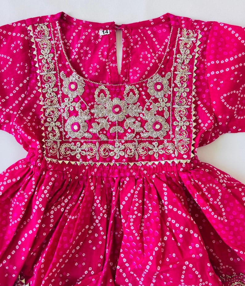 May include: A bright pink dress featuring a white tie-dye pattern and a square neckline. The bodice is decorated with silver metallic embroidery and small circular mirrors. A white tag with the number 14 is visible at the center of the neckline.