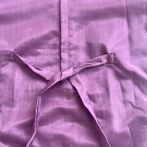 Puede incluir: Una prenda de color morado s&oacute;lido con una costura central vertical y un cintur&oacute;n de tela fino que pasa a trav&eacute;s de dos trabillas laterales. El material tiene una textura sutilmente arrugada con un ligero brillo, creando un aspecto uniforme.