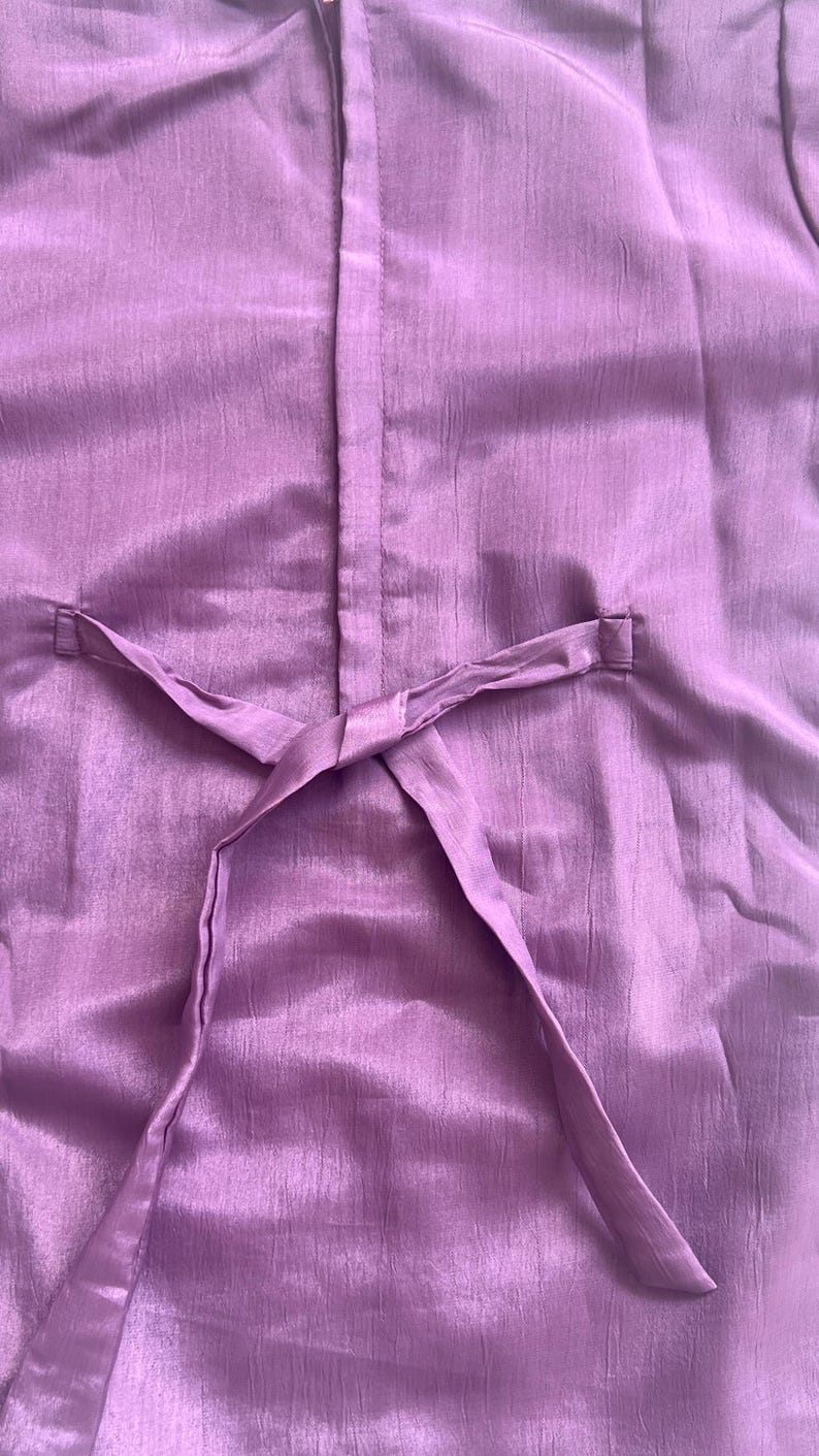 Puede incluir: Una prenda de color morado s&oacute;lido con una costura central vertical y un cintur&oacute;n de tela fino que pasa a trav&eacute;s de dos trabillas laterales. El material tiene una textura sutilmente arrugada con un ligero brillo, creando un aspecto uniforme.