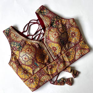 May include: A sleeveless, patterned top with a V-neckline. The top features a rich color palette of burgundy, yellow, and teal, with intricate floral and geometric designs. The back has a criss-cross tie closure. The edges are trimmed with gold detailing.