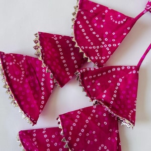 May include: A set of four fuchsia pink fabric triangles featuring a white tie-dye dot pattern. Each triangular piece is finished with a metallic gold scalloped trim along the curved edge and includes a thin matching fabric strap at the top corner.