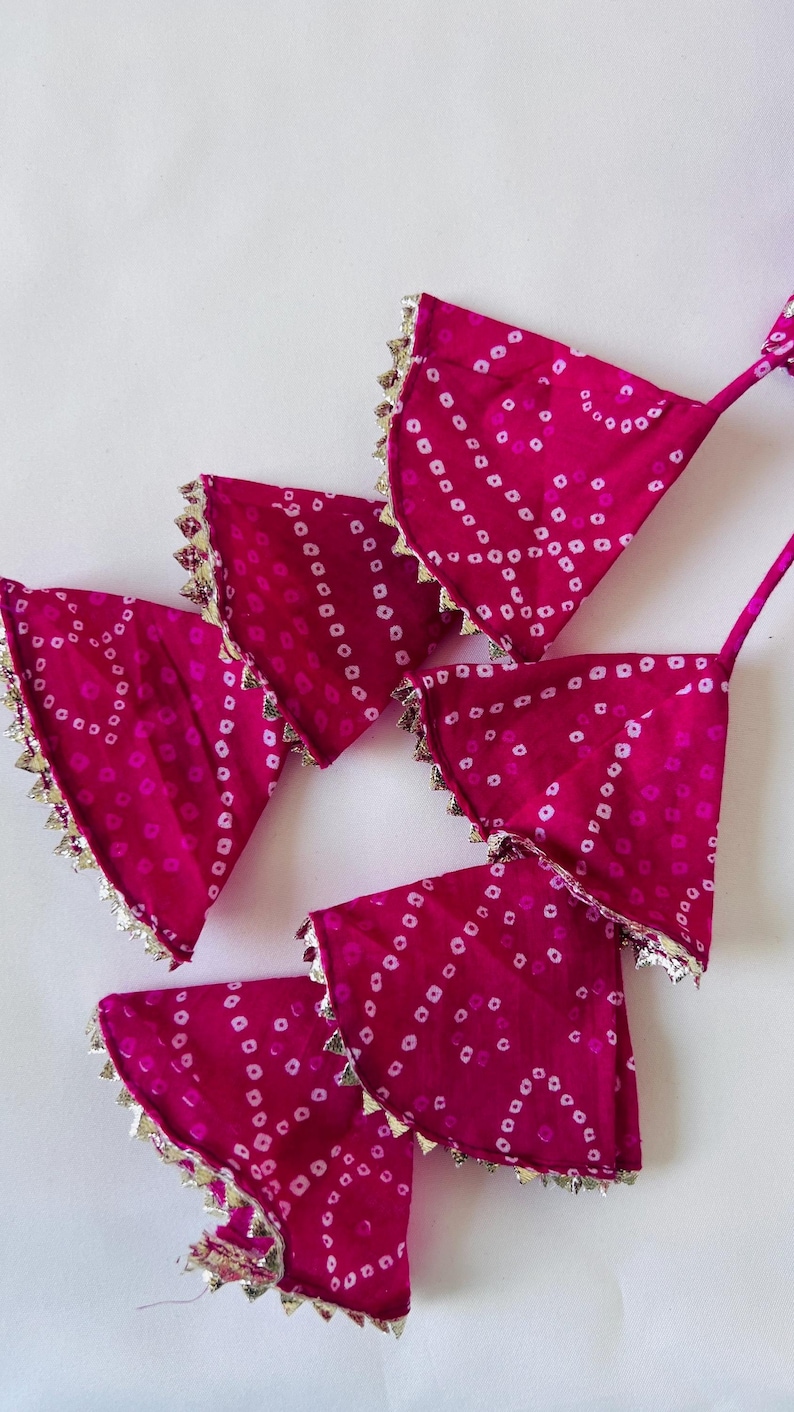 May include: A set of four fuchsia pink fabric triangles featuring a white tie-dye dot pattern. Each triangular piece is finished with a metallic gold scalloped trim along the curved edge and includes a thin matching fabric strap at the top corner.