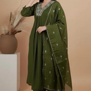 May include: Olive green three-piece ensemble featuring a long-sleeved tunic with a V-neckline and intricate embroidery, matching pants, and a sheer scarf. The outfit is adorned with delicate white floral embroidery and is paired with heeled sandals.