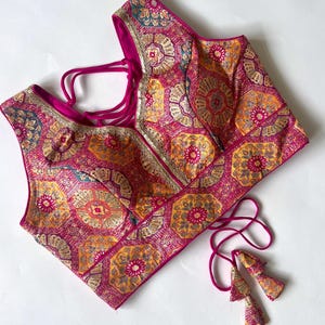 Puede incluir: Una blusa sin mangas con un estampado en tonos rosa, naranja y dorado. El diseño intrincado presenta motivos florales y geométricos. La blusa tiene un ribete rosa oscuro y lazos, con borlas a juego.