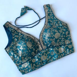 May include: Teal-colored blouse with a V-neck and floral pattern. The blouse has gold accents and a decorative trim along the neckline. A matching tie is attached at the back. The garment is made of a shiny fabric.