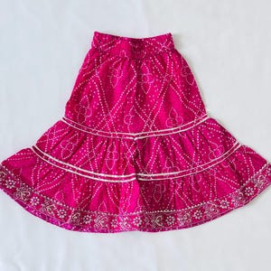 May include: A vibrant magenta skirt featuring a white dotted pattern and a tiered design. The skirt has an elasticized waistband and two horizontal rows of silver metallic trim, with a decorative floral border along the bottom hemline.