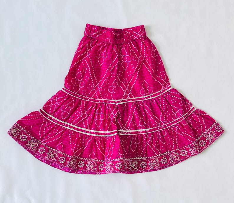 May include: A vibrant magenta skirt featuring a white dotted pattern and a tiered design. The skirt has an elasticized waistband and two horizontal rows of silver metallic trim, with a decorative floral border along the bottom hemline.