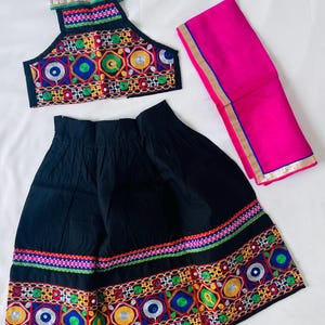 May include: A traditional Indian outfit featuring a black skirt and halter top, both adorned with colorful embroidered patterns. A bright pink scarf with gold trim completes the ensemble. The embroidery includes blue, green, yellow, and red accents.