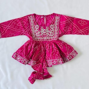 May include: A vibrant fuchsia long-sleeved peplum top featuring a white tie-dye pattern and intricate silver embroidery around the round neckline, cuffs, and hem. The garment includes a central front tie detail and a keyhole button closure.
