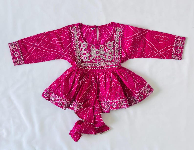 May include: A vibrant fuchsia long-sleeved peplum top featuring a white tie-dye pattern and intricate silver embroidery around the round neckline, cuffs, and hem. The garment includes a central front tie detail and a keyhole button closure.