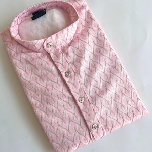 Puede incluir: Una camisa de manga larga de color rosa claro con cuello mao y un patrón de textura repetitivo en forma de chevrón blanco. La camisa tiene una tapeta frontal con cuatro botones redondos visibles y una etiqueta oscura en el cuello.