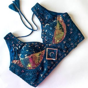 May include: A teal-colored, embroidered top with intricate floral patterns and sequin embellishments. The top features a square sequined detail and a tie closure. The design incorporates a mix of textures and colors, creating a visually rich garment.