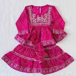 May include: A vibrant fuchsia long-sleeved tunic and tiered skirt set featuring a white tie-dye pattern. The tunic has a round neckline with intricate silver embroidery and a keyhole back closure. The skirt has silver trim along the tiers.
