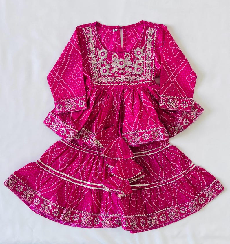May include: A vibrant fuchsia long-sleeved tunic and tiered skirt set featuring a white tie-dye pattern. The tunic has a round neckline with intricate silver embroidery and a keyhole back closure. The skirt has silver trim along the tiers.